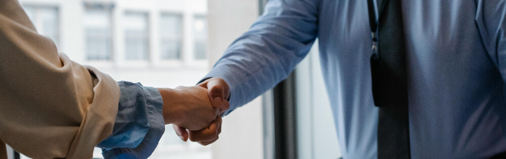 Kautionsversicherung Zwei Personen im formellen Outfit schütteln sich die Hand, im Hintergrund ein Fenster mit verschwommener Aussicht.
