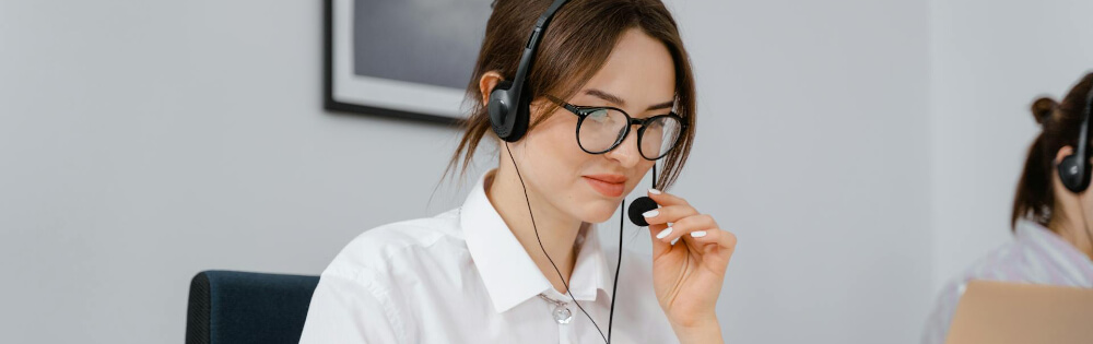 Lebensarbeitszeitkonten Eine Frau mit Brille und Headset sitzt an einem Schreibtisch und spricht in ein Mikrofon. Im Hintergrund ist eine andere Person zu sehen, die an einem Laptop arbeitet.