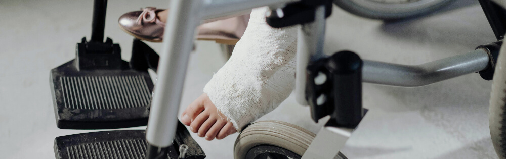 Krankentagegeld Nahaufnahme eines Fußes in einem Gipsverband, der auf der Fußstütze eines Rollstuhls ruht. Der andere Fuß trägt einen rosa Schuh. Der Rollstuhl hat schwarze Fußstützen und silberne Rahmen.